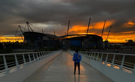 A Manchester City fan stands on a bridge leading to the club's home ground, the Etihad Stadium, behind which the sun is setting.