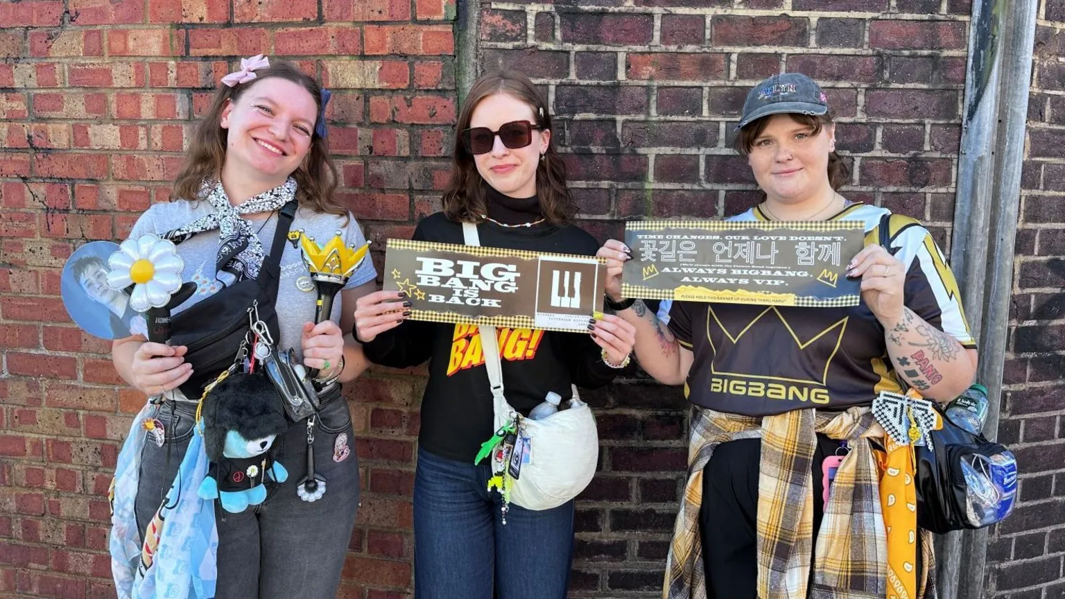 Three women standing in front of a brick wall display concert merchandise and banners. One holds a flower-shaped light stick and a crown-shaped light stick, with a cross-body bag, keyrings and a blue shopping bag. The centre woman holds a banner reading “BIG BANG IS BACK” and carries a white shoulder bag. The third holds a banner with Korean and English text and wears a clear waist bag. Various accessories, badges and merchandise are attached to bags and clothing.