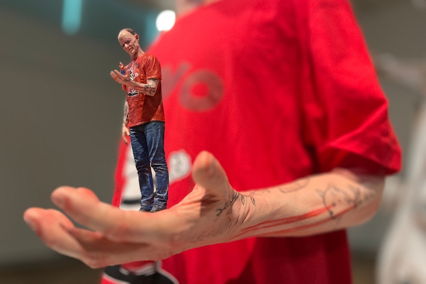 A sculpture of a man in a red t-shirt holding a tiny of himself in his palm, who is holding a tinier version in its palm.