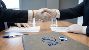 Manos enguantadas de dos personas dándose la mano sobre una mesa. Se observan una carpeta con el logo del BCIE, un bolígrafo azul y un pin de la bandera de El Salvador.