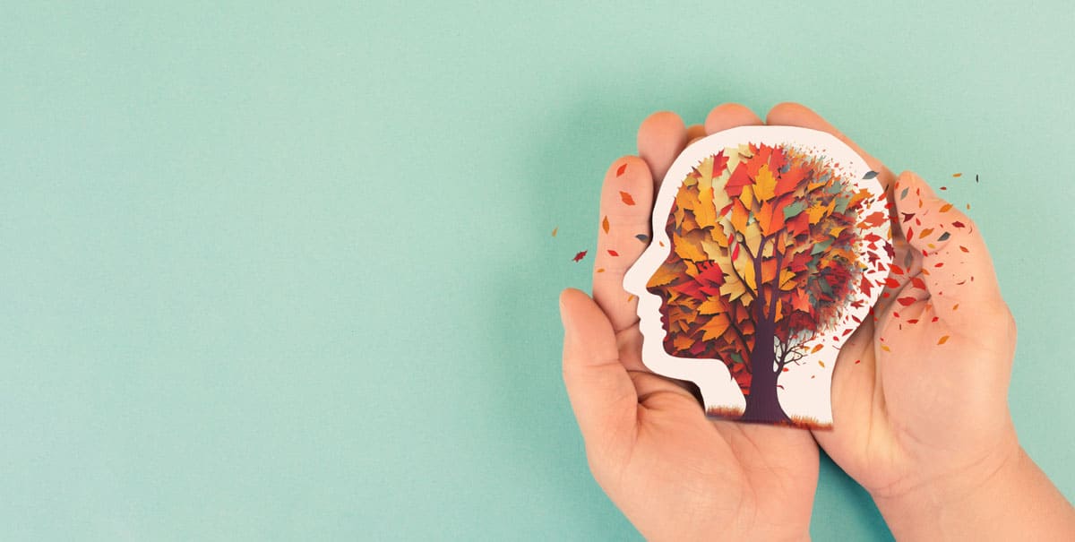 uma mão segurando um desenho de uma cabeça com o cérebro representado por uma árvore sob o vento para demonstrar as condições de um paciente com Alzheimer.