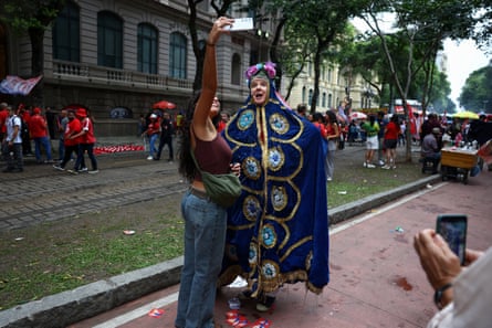 A woman takes a selfie with a Lula supporter who is dressed as Our Lady of Aparecida in a highly decorated blue robe and headdress. They are standing in a street with others gathered behind them.